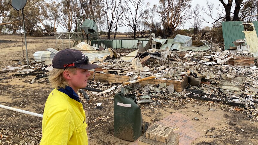 A young man, wearing a high viz shirt, stands in front of a house destroyed by fire