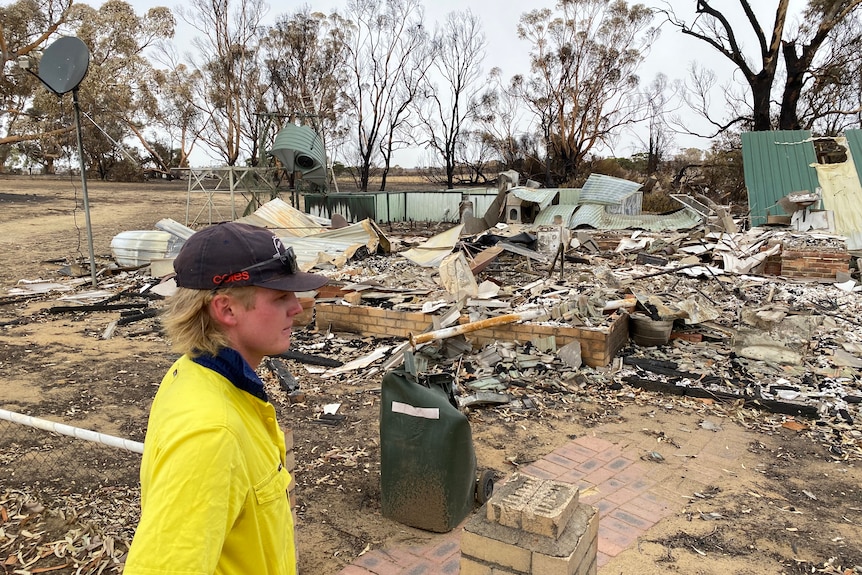 A young man, wearing a high viz shirt, stands in front of a house destroyed by fire