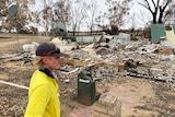 A young man, wearing a high viz shirt, stands in front of a house destroyed by fire