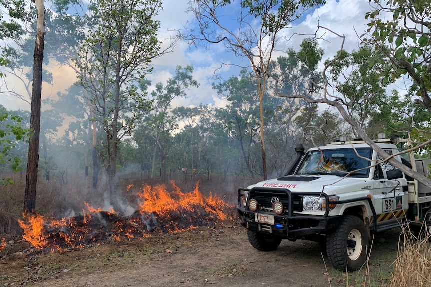 A bushfire burns amidst grass while a white ute is parked nearby.