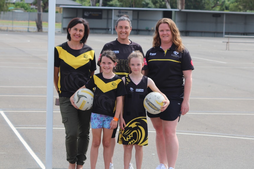 Three women and two girls stand on bitumen netball courts and smile.