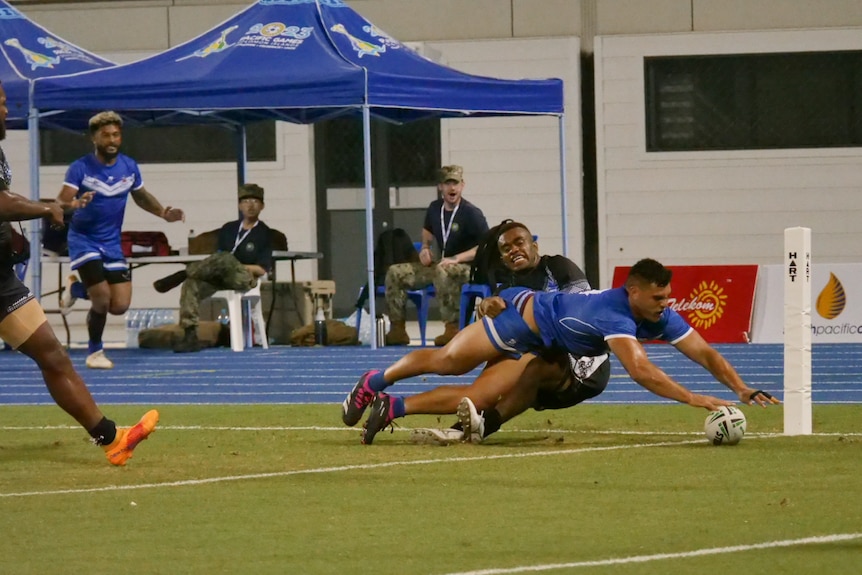A Samoan player scores a try, diving with his hand on the ball as a Fijian player tries to stop him.