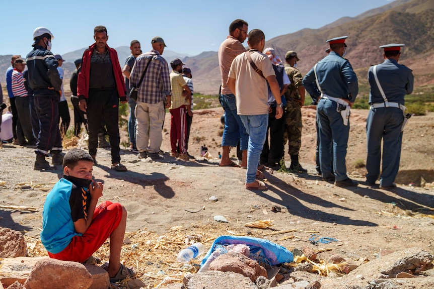 A boy sits wearing a blue shirt and red pants covering his mouth as men stand watch nearby.