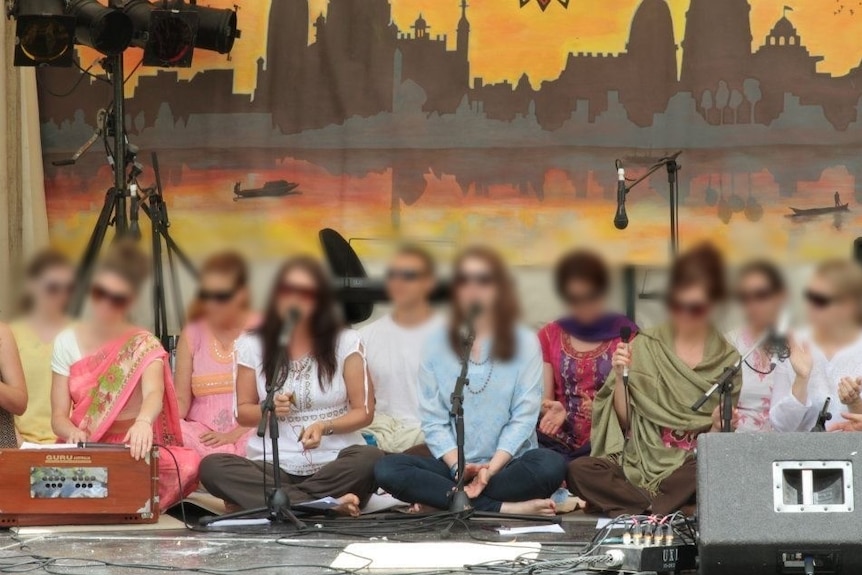 Ten people wearing sunglasses sit on a stage. Their faces are blurred. Some speak into microphones.