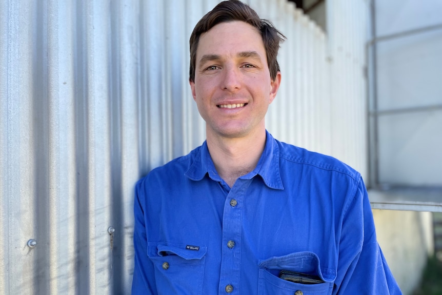 A man in a blue shirt leans beside a shed.