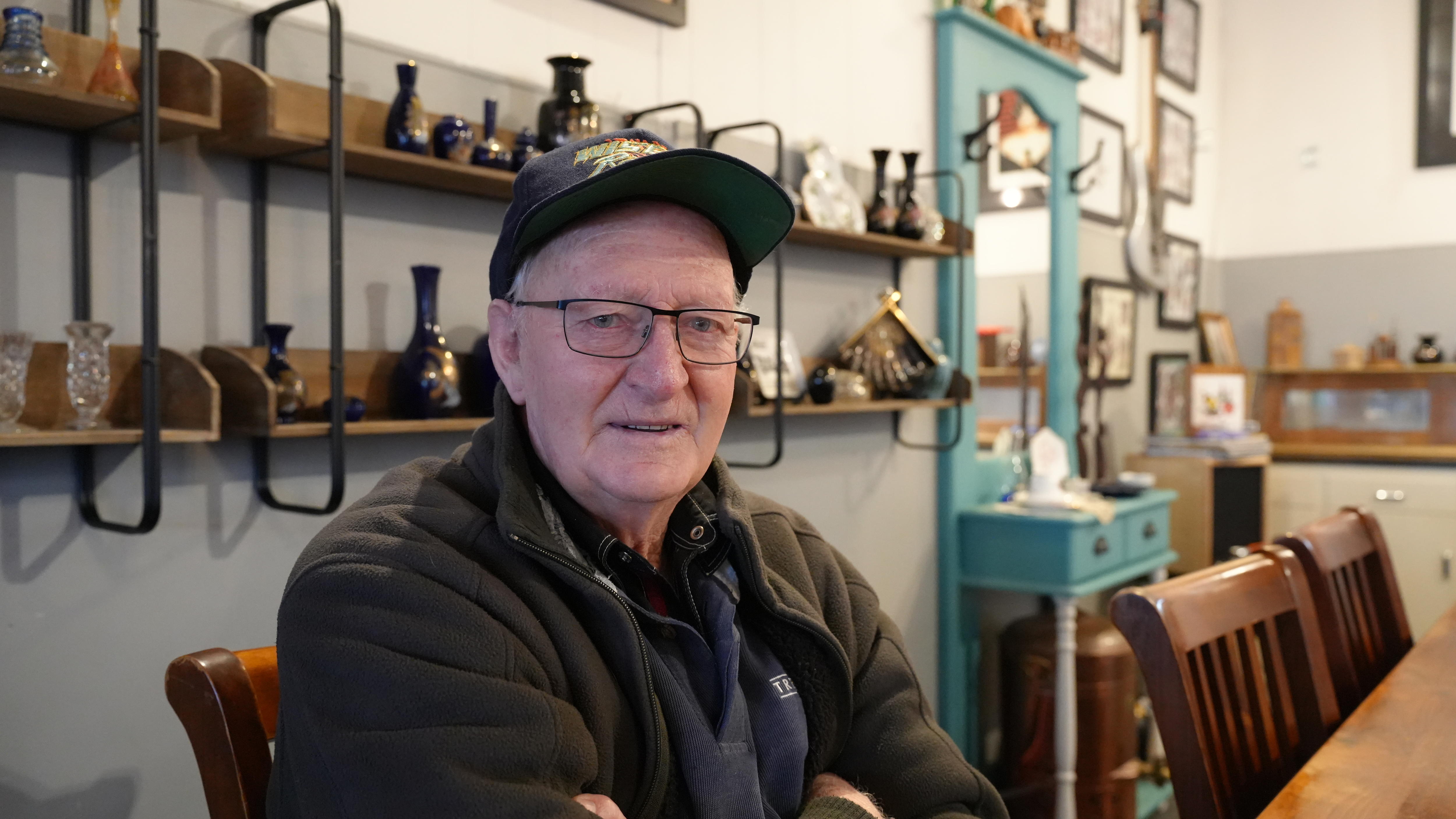 An older man in black sits with his arm folded in a cafe.