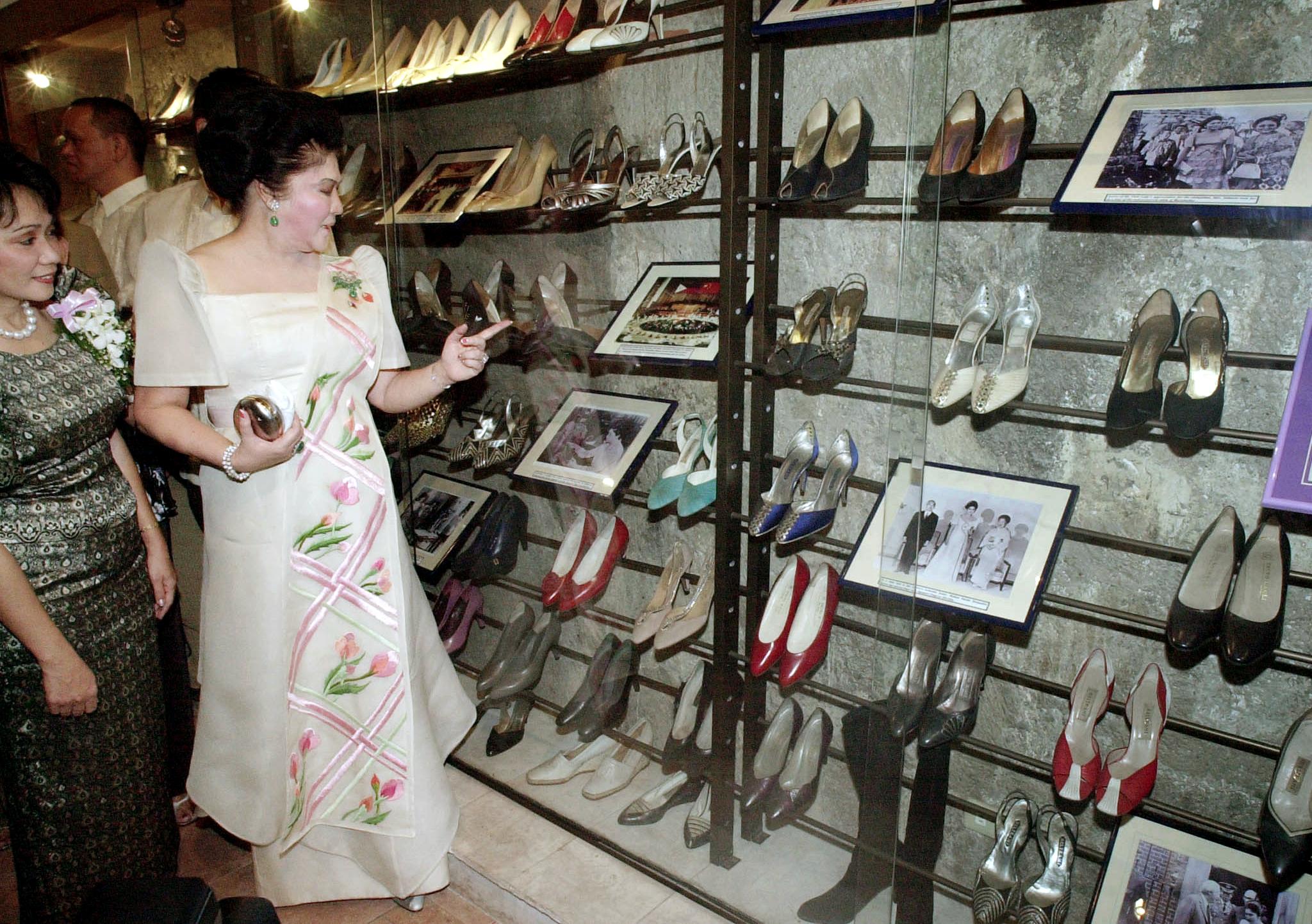 Imelda Marcos in a white babydoll dress with pink flowers gazes at her collection of designer shoes in glass cabinets