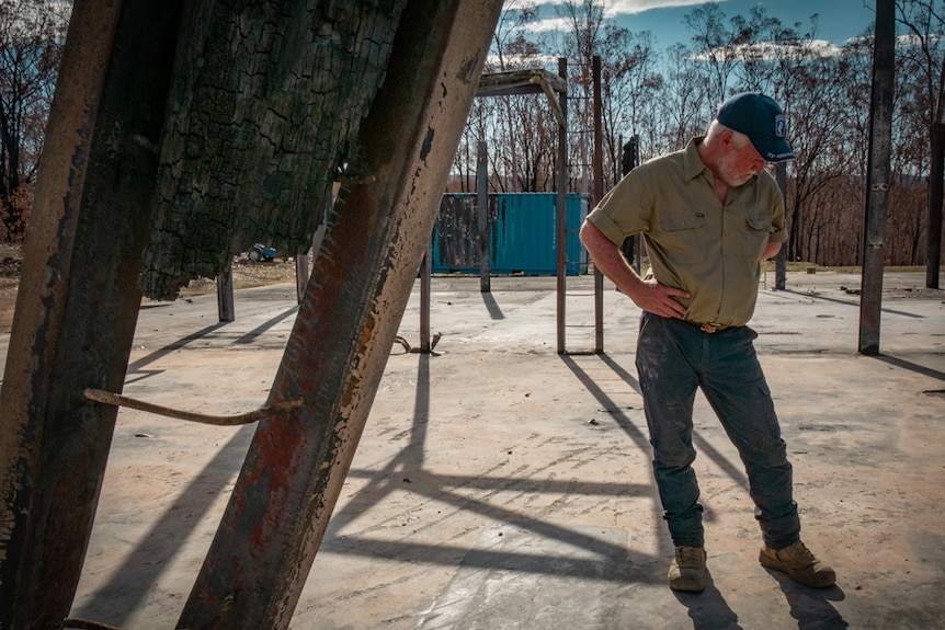 Man stands on a concrete slab with scorched and twisted girders that is all that left of his home.