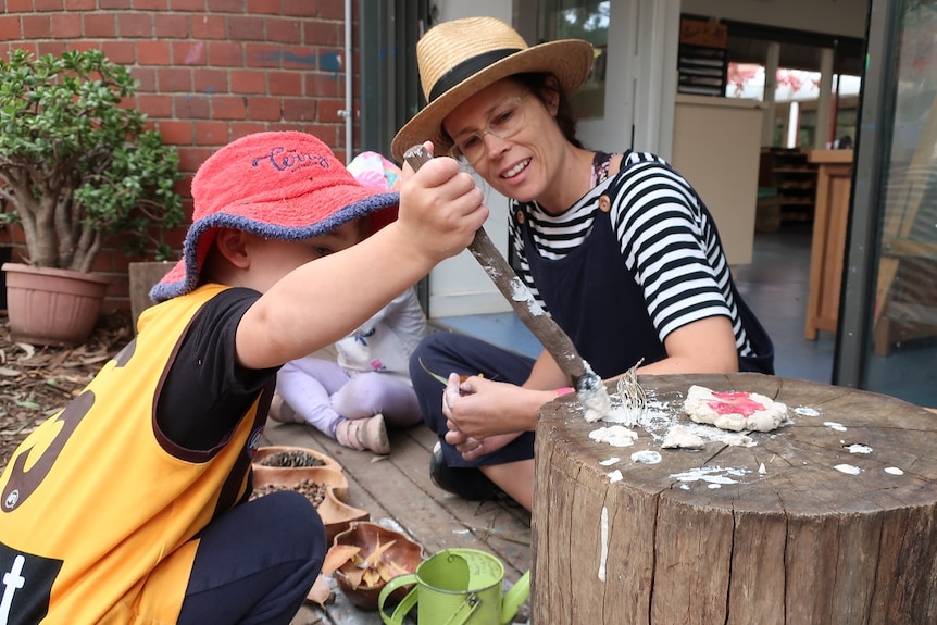 A woman wearing a straw hat watches a young boy painting with a stick.