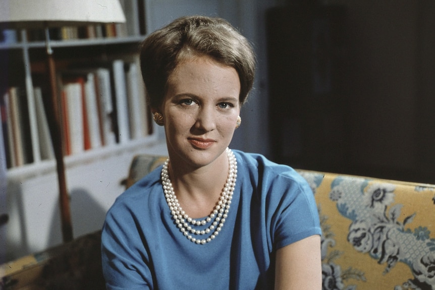 Princess Margrethe, a young woman wearing blue dress and a string of pearls around her neck, sits on a sofa
