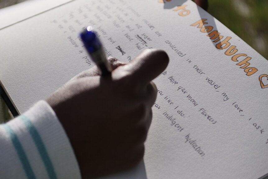 A teenage girl writes on a piece of paper. Several lines are seen under a heading "Ode to Kombucha".