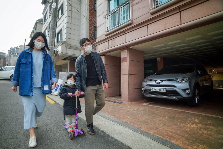 A Korean couple in face masks walk next to a small boy on a razor scooter 