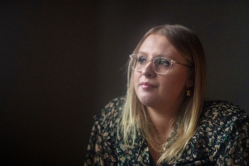 A blonde woman wearing a floral blouse and glasses looks into the camera in soft window light and dark background.