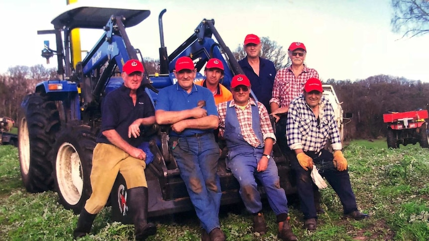 A group of volunteers wearing red caps and work clothes lean up against an earth mover.