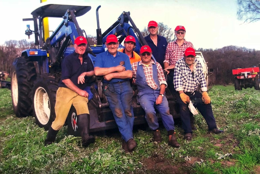 A group of volunteers wearing red caps and work clothes lean up against an earth mover.