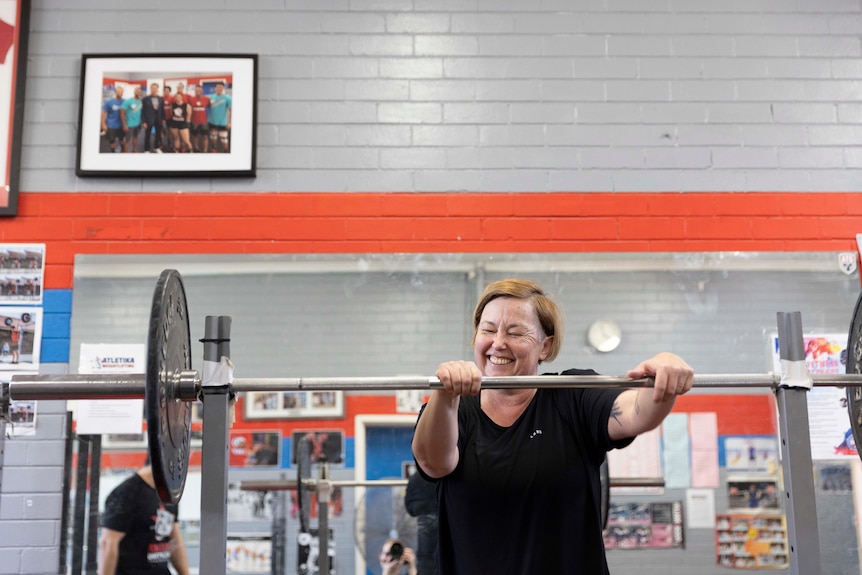 A middle-aged woman leans on a weightlifting bar and laughs