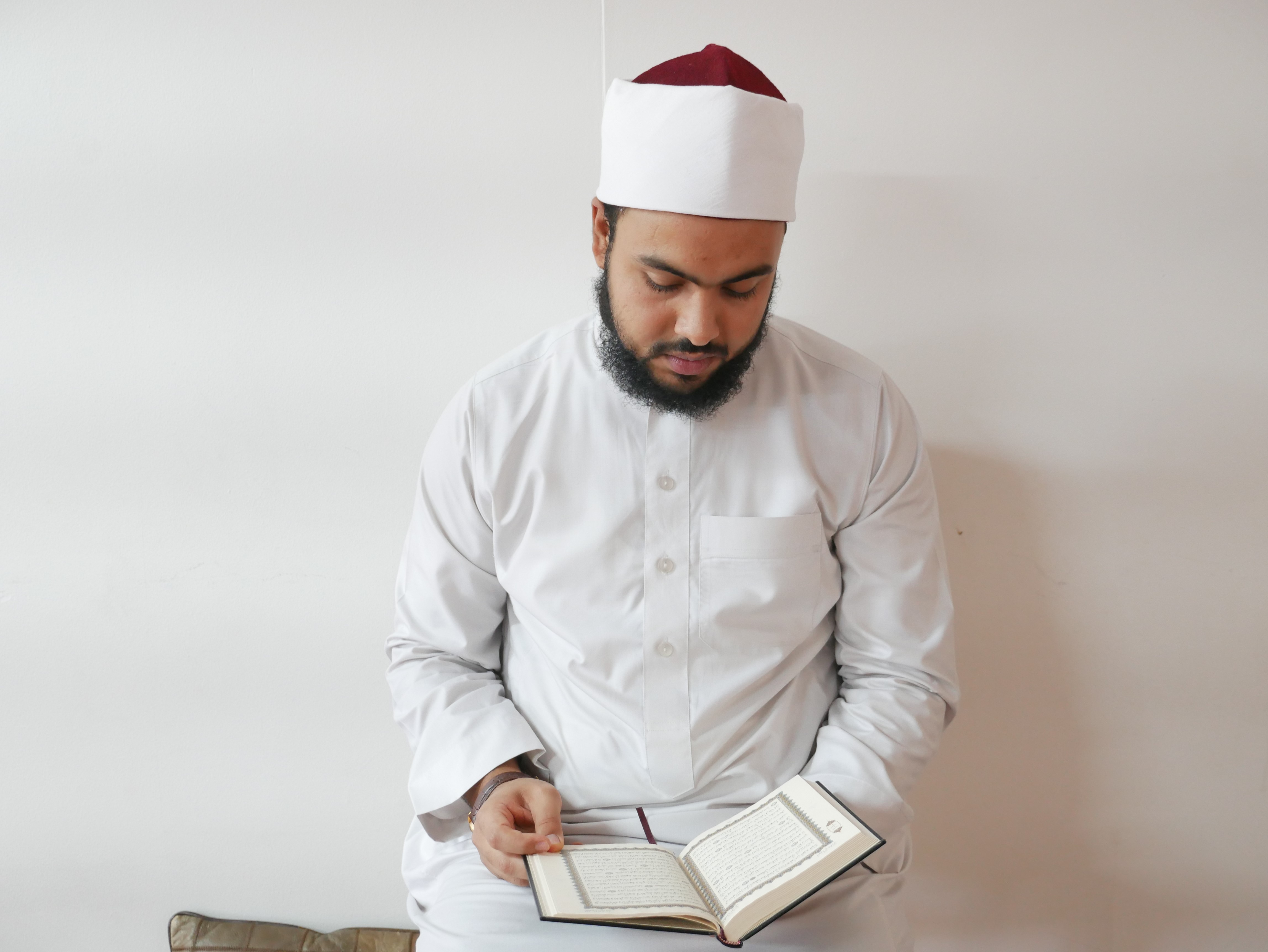 Imam Ahmad Aboukhadra sits against a plan white wall reading a book in his lap.