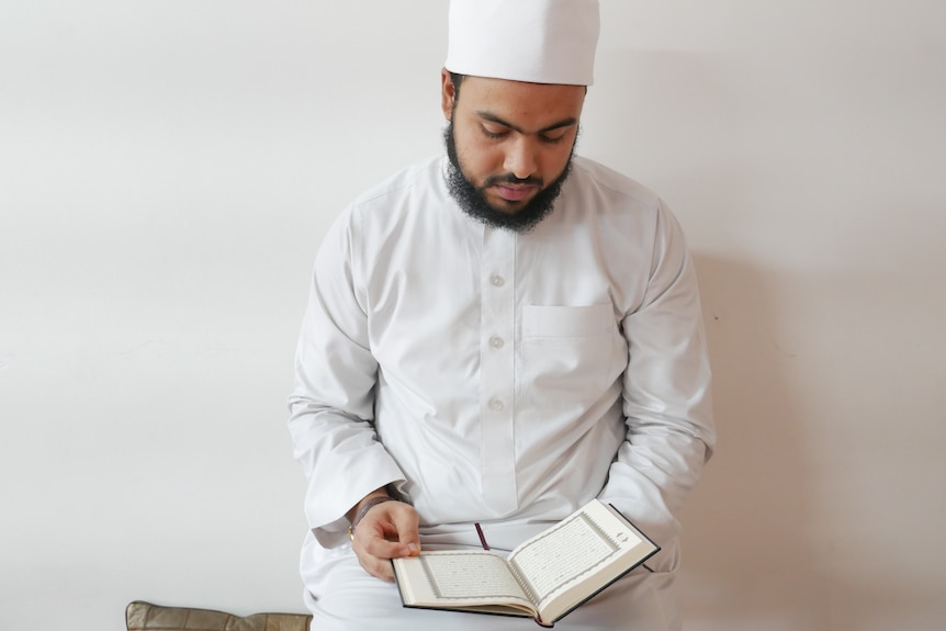 Imam Ahmad Aboukhadra sits against a plan white wall reading a book in his lap.
