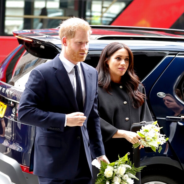 Prince Harry and Meghan Markle walk in front of a car carrying bunches of flowers