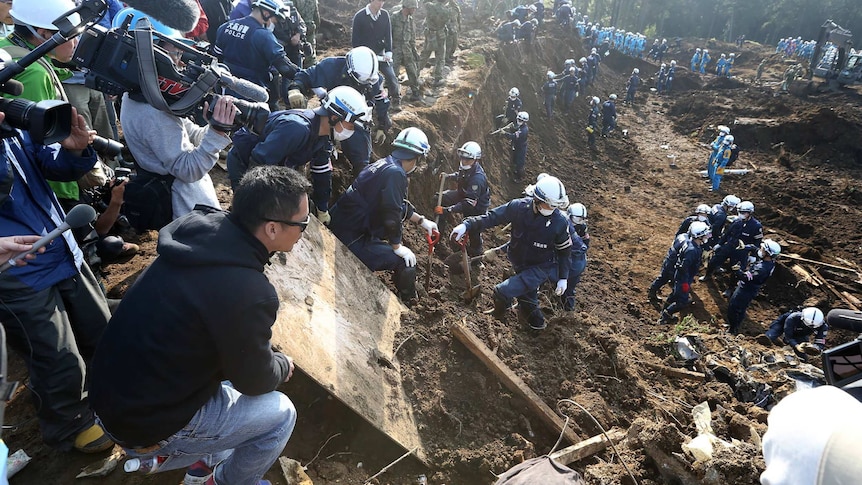 Relatives and journalists watch police officers search for missing victims.