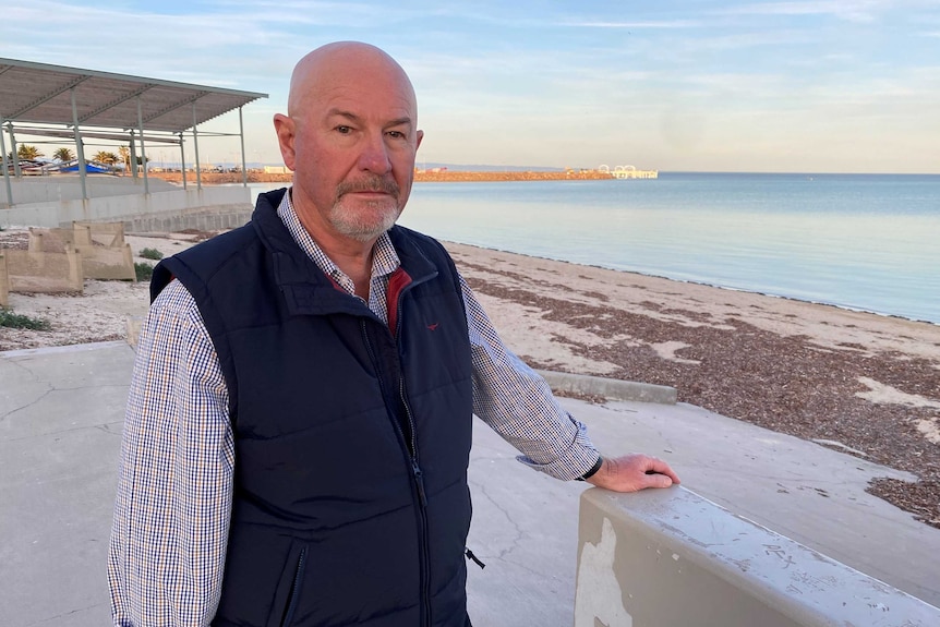 A man stands in front of a boat ramp at the beach. There is a long pier behind him.