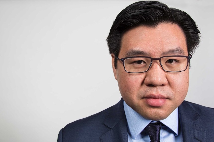 Tim Soutphommasane stands against a white wall dressed in a suit and tie.