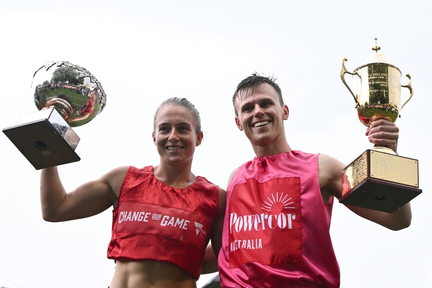 A young woman and man in red and pink shirts with fair skin hold up golden trophies.