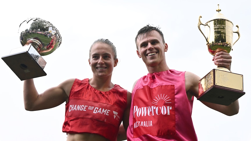 A young woman and man in red and pink shirts with fair skin hold up golden trophies.