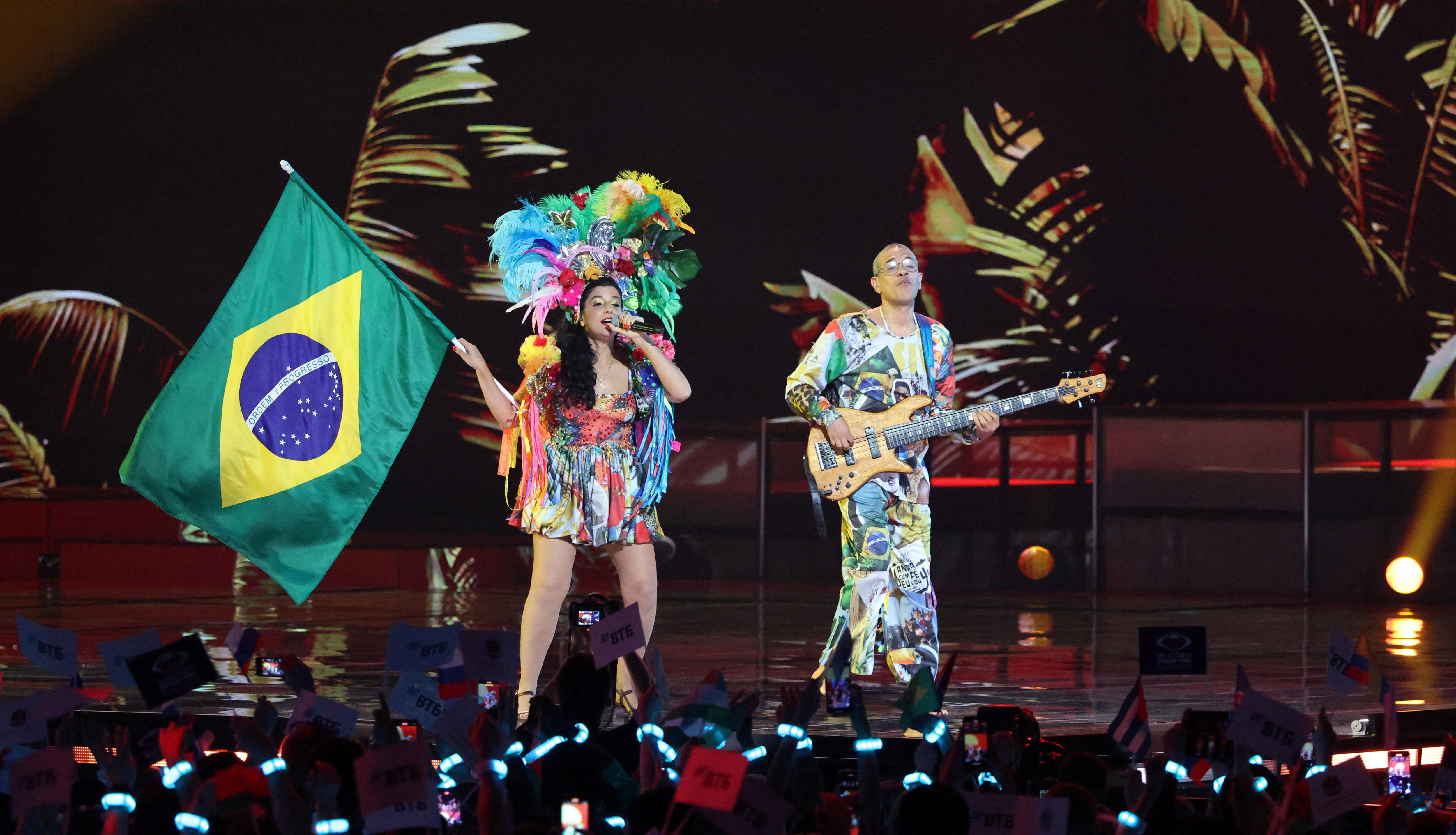 A woman in a bright dress and headress holds a Brazilian flag and sings on a stage next to a man playing a bass guitar