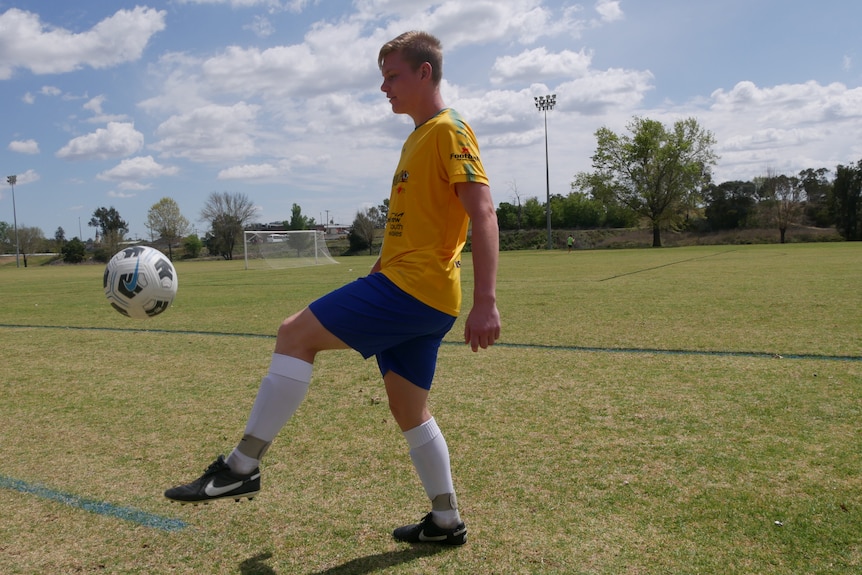 A young man juggles a soccer ball with a yellow jersey on.