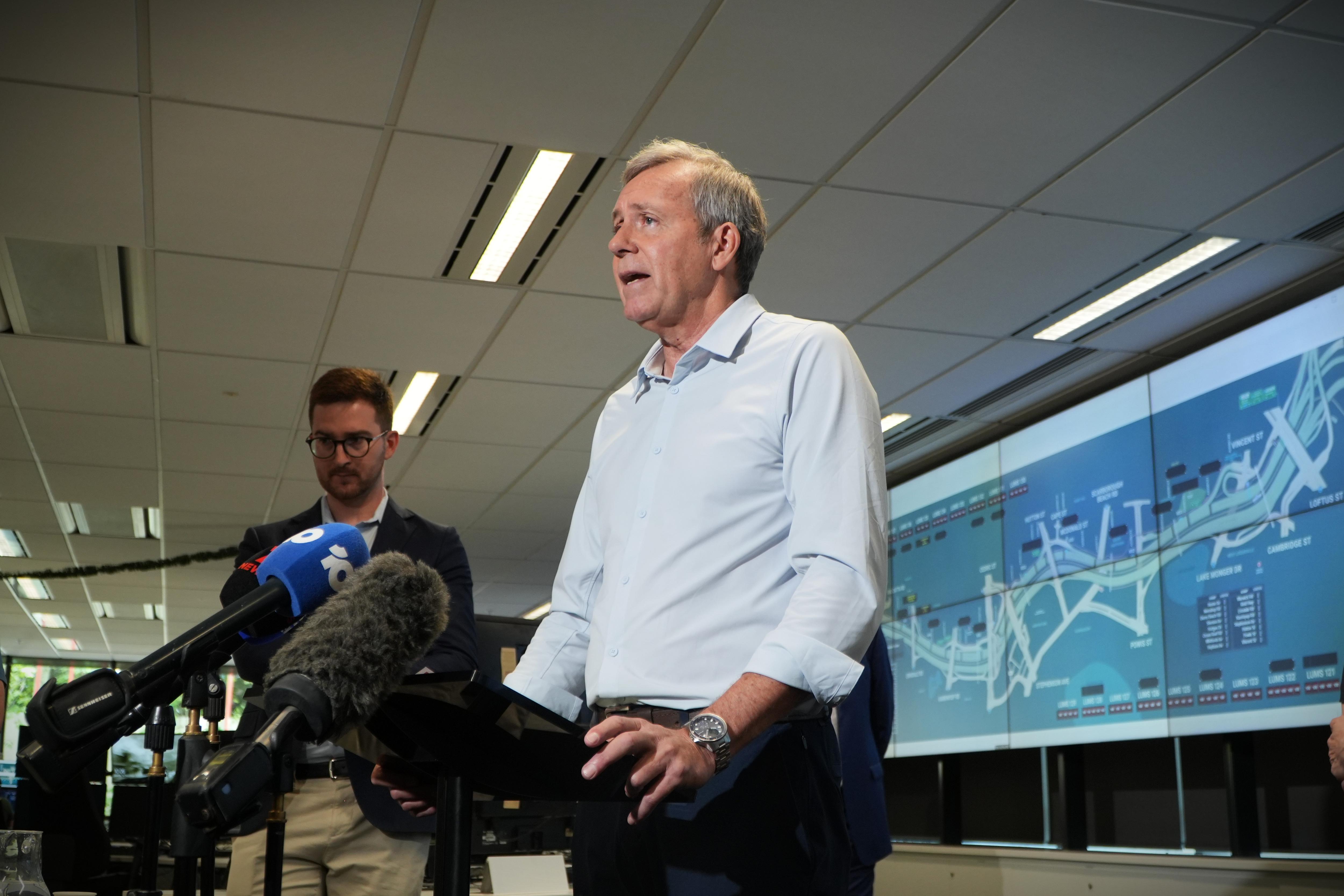 Reece Whitby fala em uma coletiva de imprensa com telas e outro homem visível ao fundo. 