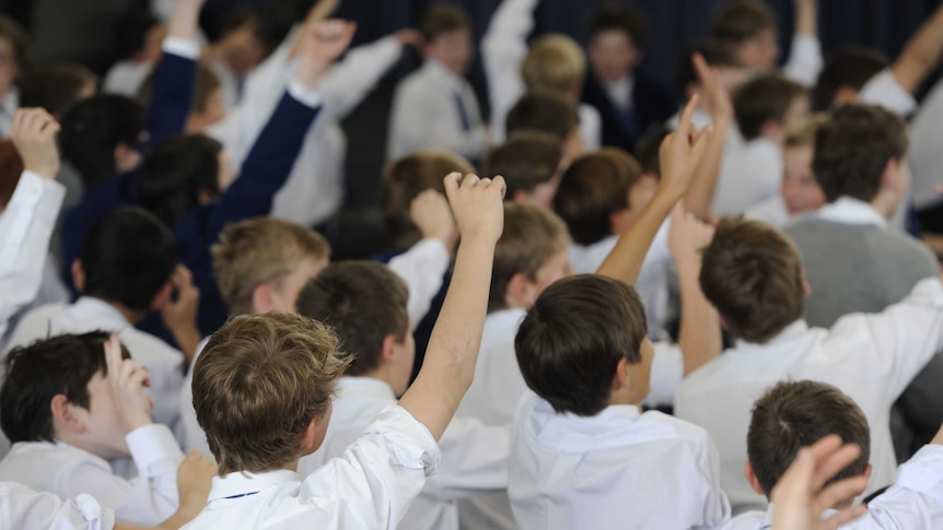 Students sit during a talk at a private college with their hands in the air wearing white shirts.