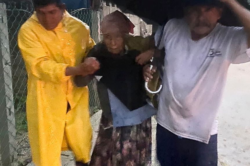 Двое мужчин помогают пожилой женщине на улице.  Они держат зонтик и, кажется, бегут.