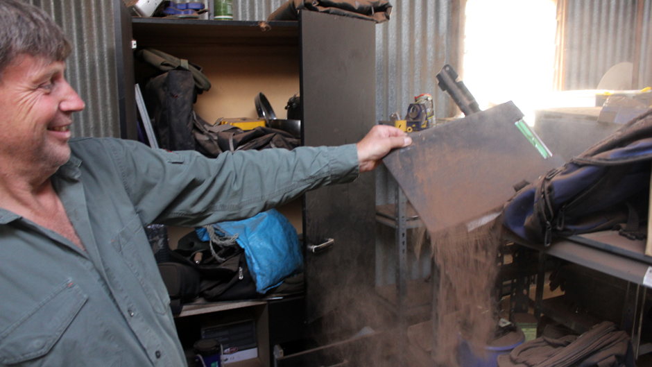 A man in a green shirt and jeans brushes red dust off a clip board in a tool shed.