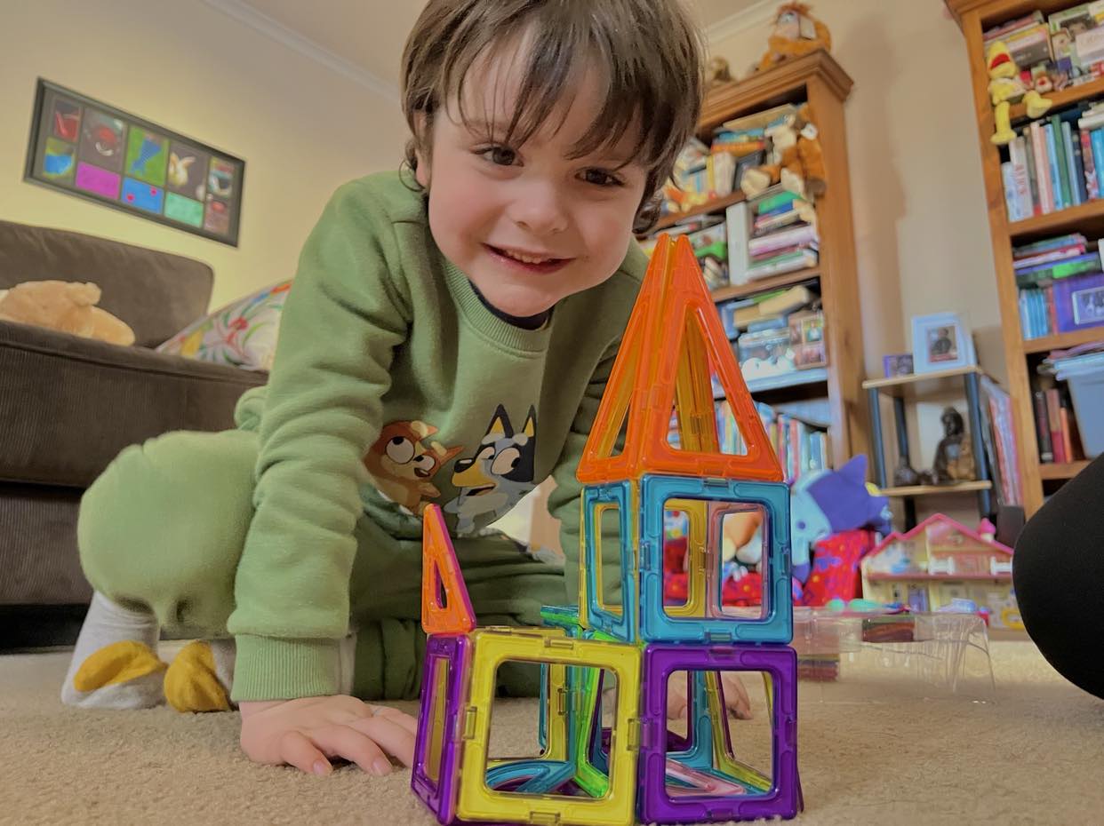 Little boy playing with blocks and smiling, in front of two full boo shelves and other toys