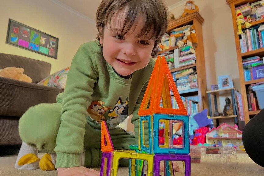 Little boy playing with blocks and smiling, in front of two full boo shelves and other toys