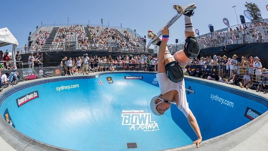 A young girl wearing a helmet and knee pads, hanging upside down on the edge of a skate bowl.