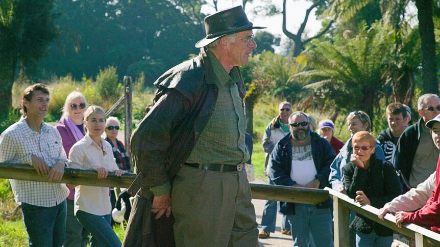 A man in a drizabone and akubra addresses a crowd of tourists in an outdoor forest setting.
