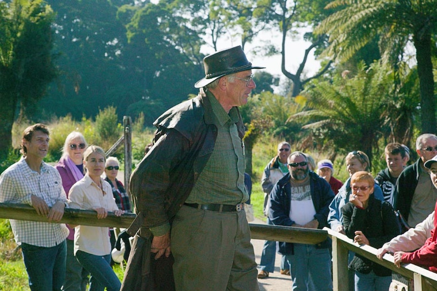 A man in a drizabone and akubra addresses a crowd of tourists in an outdoor forest setting.