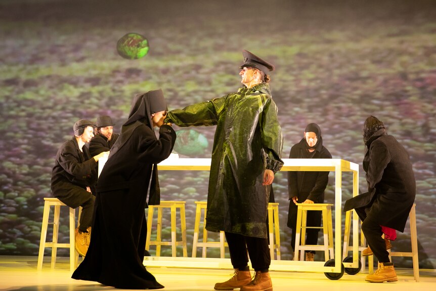 An actor in all-black kisses the hand of another actor in a green coat and military hat on a stage. Four actors sit behind them