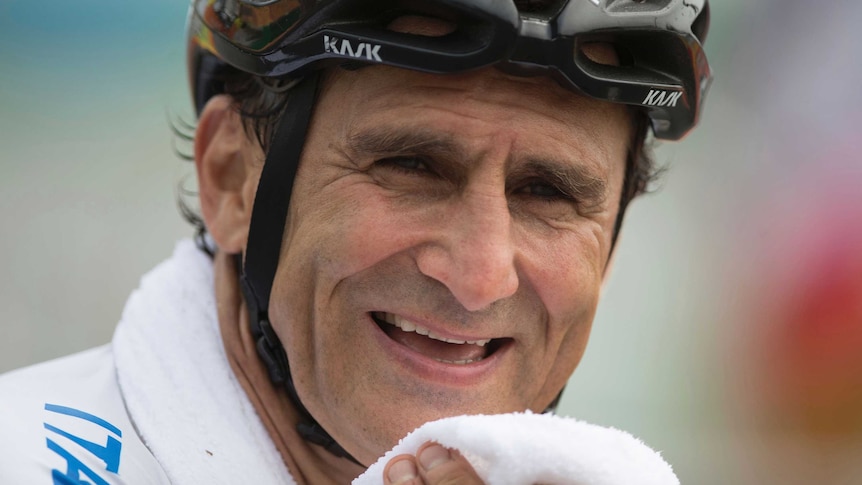 Alex Zanardi holds a towel to his smiling face while wearing a bike helmet