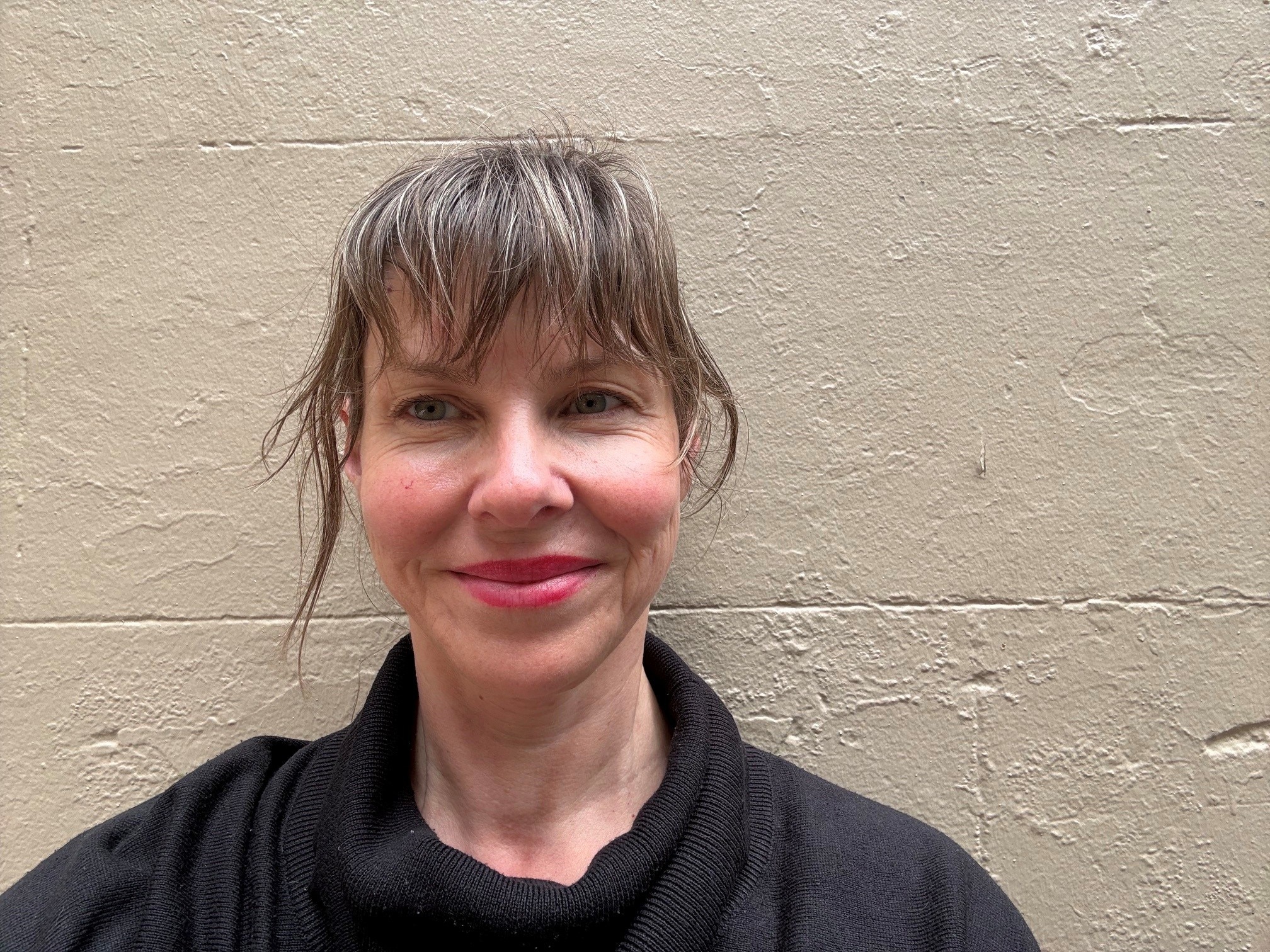 A woman wearing black and with bangs smiles at the camera, with a cream wall in the background