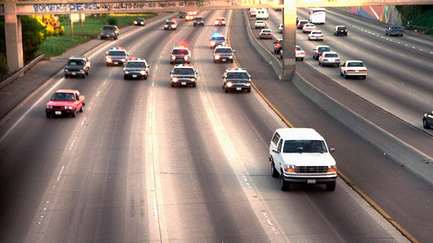 A view of a highway with a white SUV in the front and several police cars with sirens behind