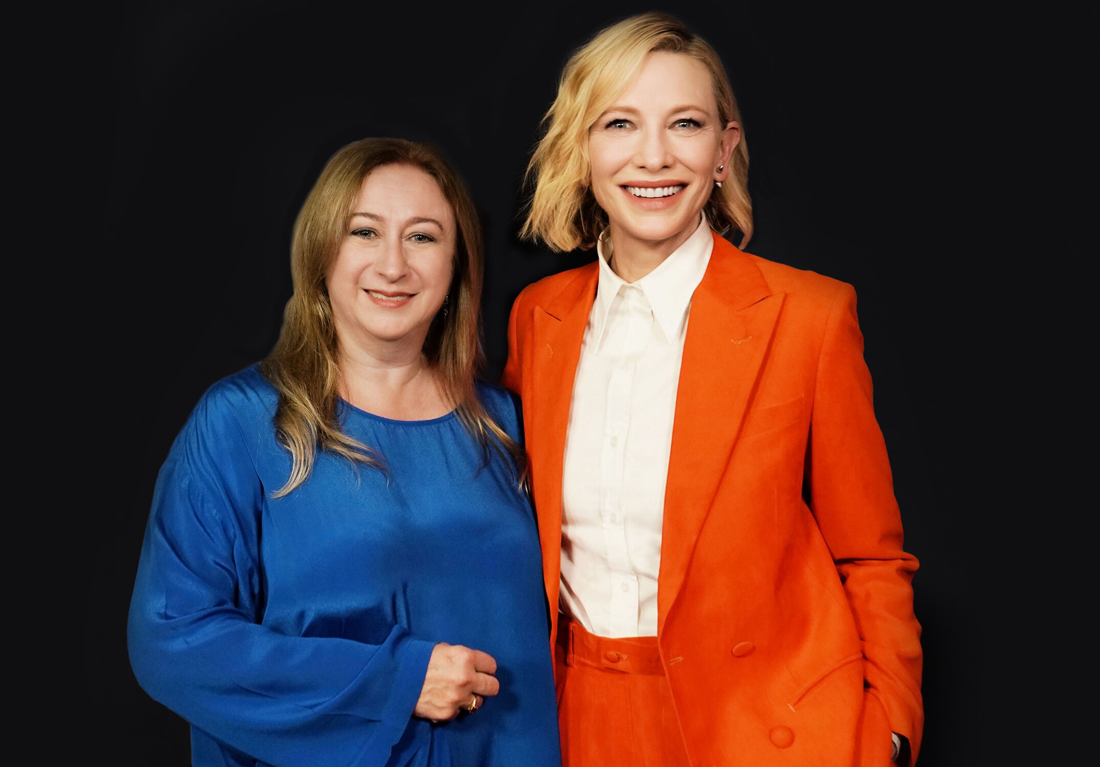 Two middle-aged women, one with long brown hair, the other with short blonde hair, wearing an orange suit, smile brightly