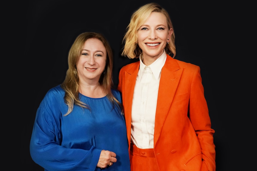 Two middle-aged women, one with long brown hair, the other with short blonde hair, wearing an orange suit, smile brightly
