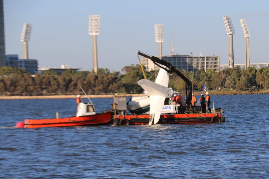 The torn-off tail of a plane is lifted onto a barge with the lights from the WACA Ground in the background.
