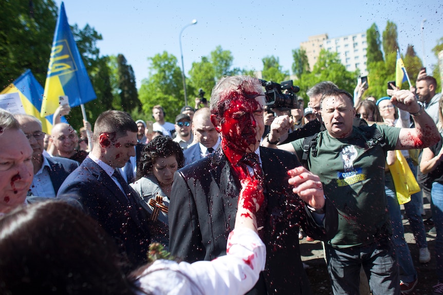 Man in a suit, walking through a crowd, has red paint thrown in his face.