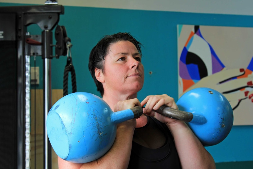 A woman holding two large, heavy kettlebells up to her chin