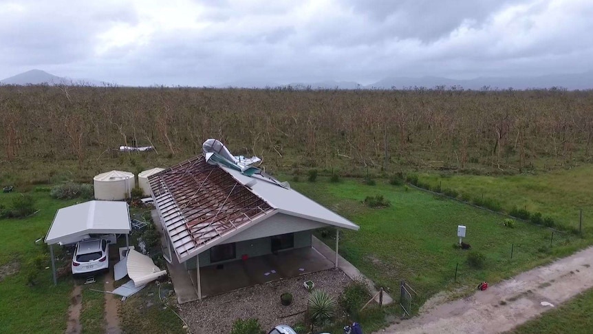 Drone footage of a green and grey house in a field with its tin roof ripped off.