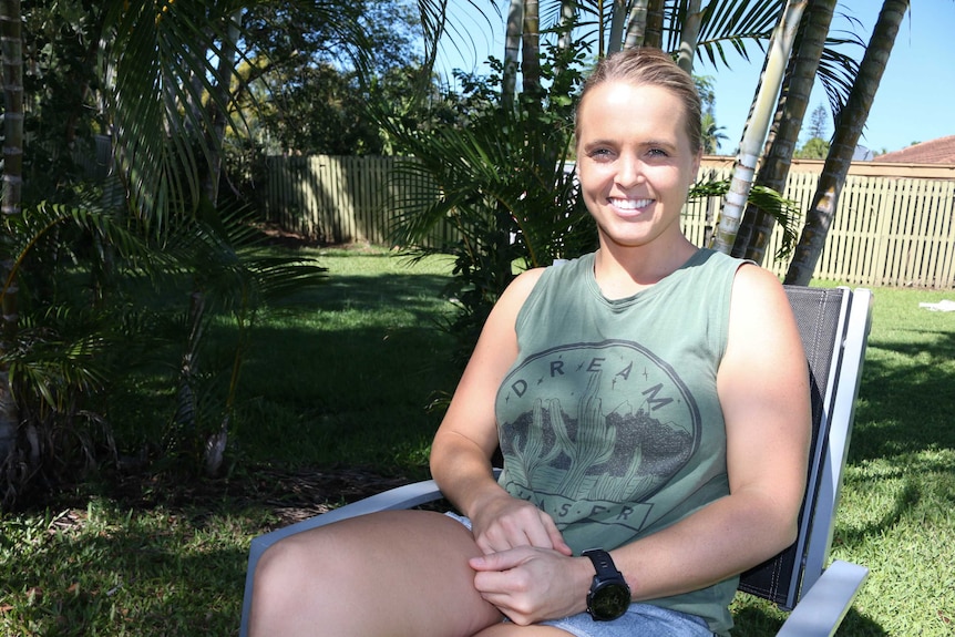 Woman with blonde hair sitting on chair in back yard with golden cane palm trees behind her.